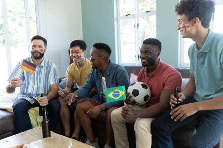 Happy multiracial male friends with flags, beer bottles, balls watching match while sitting on sofa....