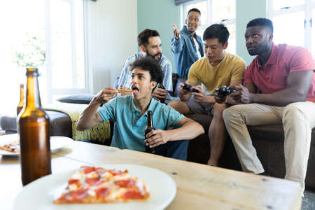 Multiracial male friends playing video game and enjoying beer and pizza in living room, copy space....