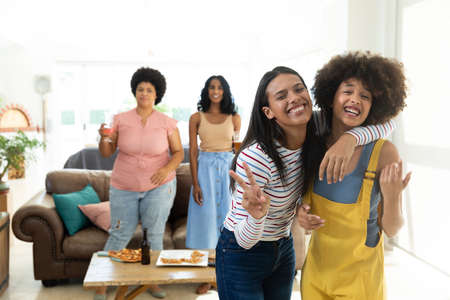 Happy biracial young woman showing peace sign while enjoying with friends at home, copy space. Unaltered, friendship, togetherness, social gathering, enjoyment and weekend activities concept.の写真素材