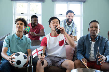 Happy multiracial male friends with ball and beer bottles watching soccer match on sofa at home. Sport, alcohol, unaltered, togetherness, social gathering, enjoyment and weekend activities.の写真素材