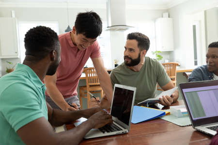 Smiling multiracial male entrepreneur talking with colleagues working at desk in office. Laptop,...