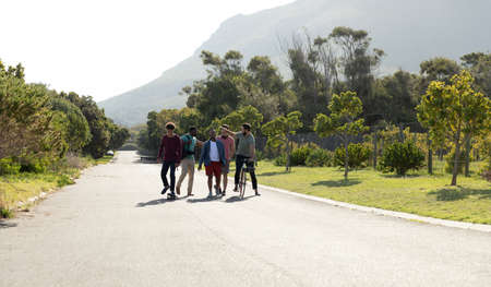 Male multiracial friends with bicycle walking on road amidst lush trees against clear sky in summer. Copy space, nature, unaltered, friendship, togetherness, getaway, holiday and weekend activities.の写真素材