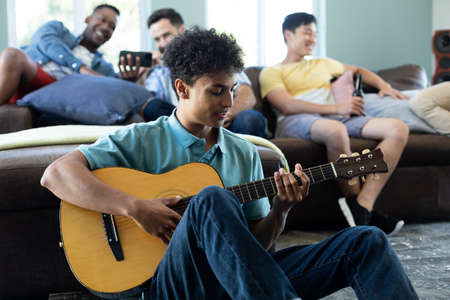 Multiracial young man playing guitar while male friends relaxing in background on sofa at home....