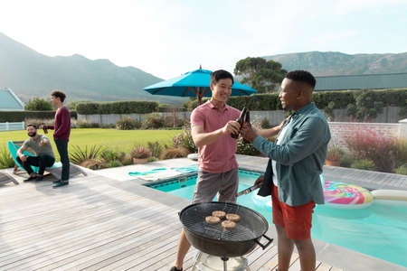 Multiracial male friends toasting beer while standing by barbecue grill in yard against sky. Copy...