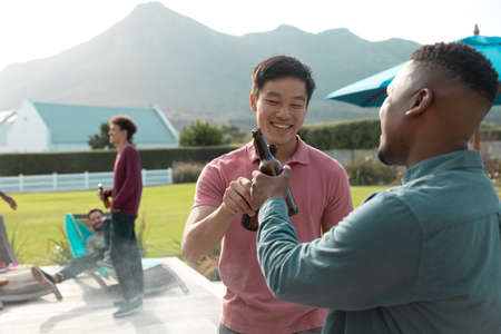 Cheerful multiracial male friends toasting beer bottle wile enjoying party in yard against mountain....