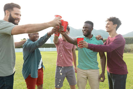 Carefree multiracial male friends toasting beer glasses while standing on grassy land in yard. Copy...