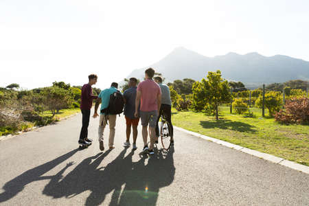 Multiracial male friends walking on road amidst plants against clear sky in summer during weekend....