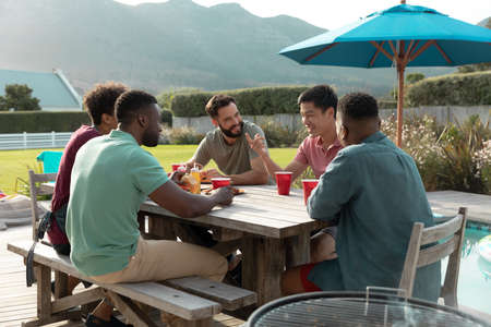 Multiracial happy male friends with drinks sitting at table and chatting in yard during summer....