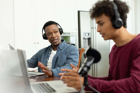 Multiracial male coworkers with headphones and laptop discussing while recording podcast in office....