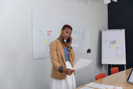 African american young businesswoman talking on smart phone and analyzing document in office. Unaltered, creative business, workplace, copy space, office, strategy, infographic, communication.の写真素材