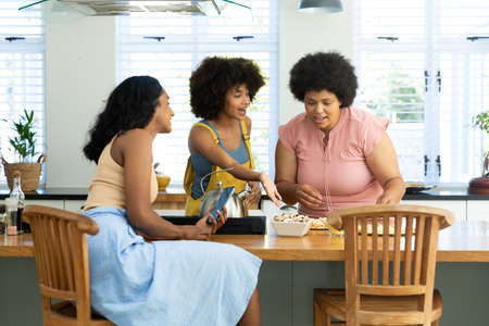 Biracial woman with smartphone looking at friends with afro hair making pizza at kitchen island....