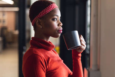 Thoughtful african american businesswoman having coffee while looking away in creative office. Unaltered, creative business, workplace, coffee break, refreshment, contemplation.の写真素材
