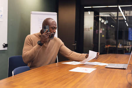 African american mature businessman examining document while talking on smart phone in office....