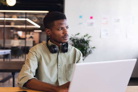 Thoughtful african american young businessman with laptop in office. Unaltered, creative business, workplace, wireless technology, contemplation.の写真素材