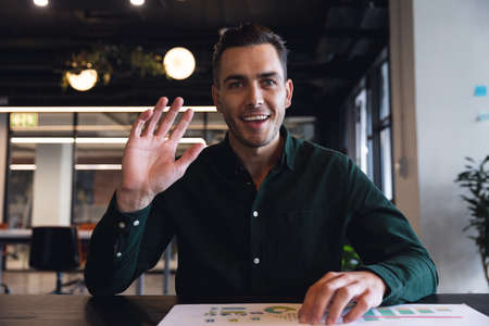 Portrait of caucasian young businessman waving hand on video call at creative office. Unaltered,...