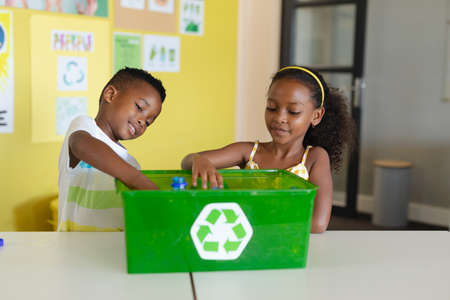 African american elementary students putting bottles in recycling container at desk in school. unaltered, education, childhood, environmental cleanup, sustainable lifestyle and school concept.の写真素材