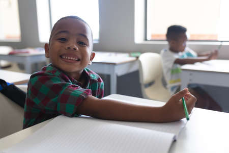 Portrait of smiling african american elementary schoolboy sitting at desk in classroom. unaltered,...