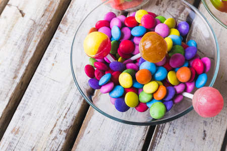High angle view of multi colored lollipops and chocolate candies in glass bowl on wooden white...