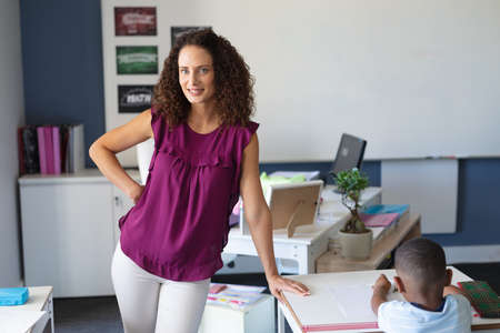 Portrait of smiling caucasian young female teacher with hand on hip standing in classroom....
