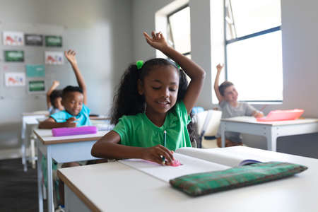 Smiling multiracial elementary school students with hands raised sitting at desk in classroom....