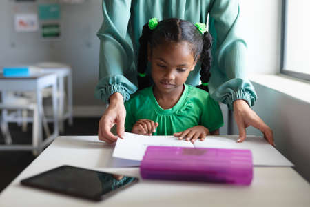 Caucasian young female teacher assisting african american elementary girl studying at desk. unaltered, education, childhood, learning, teaching, occupation and school concept.の写真素材