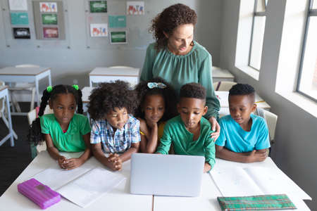 Caucasian young female teacher standing by african american elementary students using laptop at...
