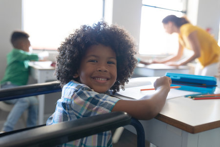 Portrait of smiling african american elementary boy sitting on wheelchair at desk in classroom....