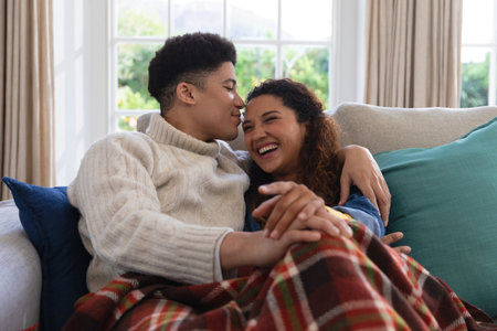 Happy biracial couple sitting on couch under blanket, laughing and embracing in living room. Inclusivity, domestic life, leisure time and togetherness concept.の写真素材