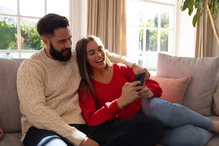 Caucasian couple sitting on couch in living room, laughing and embracing, looking at smartphone. Domestic life, leisure time, romance and togetherness concept.の写真素材