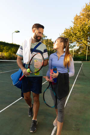 Happy caucasian couple holding rackets and water bottles talking on outdoor tennis court. Sport, healthy hobbies, competition, fitness and leisure time concept.の写真素材