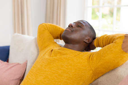 African american man lying back on couch at home with eyes closed. Inclusivity, domestic life, free time and relaxation, spending time alone at home.の写真素材