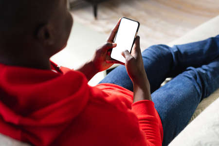 Over shoulder view of african american man lying on couch using smartphone, copy space on screen....
