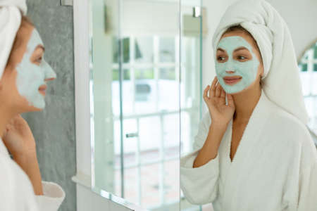Happy caucasian woman wearing robe, applying beauty face mask on face in bathroom. Spending quality...
