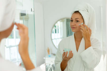 Happy caucasian woman wearing robe, applying beauty face mask on face in bathroom. Spending quality...