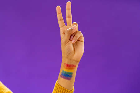 Close up of biracial man showing v sign with lgbt flag on arm on purple background. Lgbt and pride concept.の写真素材