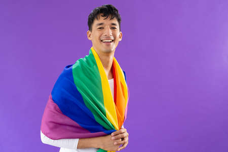 Portrait of happy biracial man holding lgbt rainbow flag and smiling on purple background. Lgbt and pride concept.の写真素材