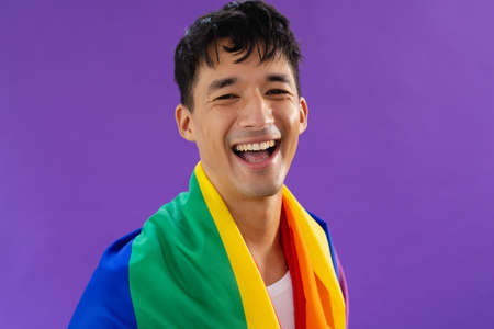 Portrait of happy biracial man holding lgbt rainbow flag and smiling on purple background. Lgbt and pride concept.の写真素材