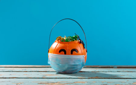 Face mask over pumpkin shaped bucket full of candies on wooden surface against blue background. fall season and celebration during covid-19 pandemic conceptの写真素材