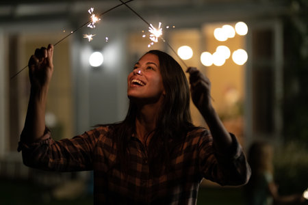 Caucasian woman holding two sparklers, smiling, outside of her home, enjoying her time. domestic...
