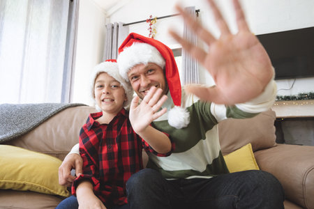 Caucasian man at home with his son at Christmas, wearing Santa hats sitting on sofa in living room, smiling and waving. Social distancing during virus quarantine lockdown.の写真素材
