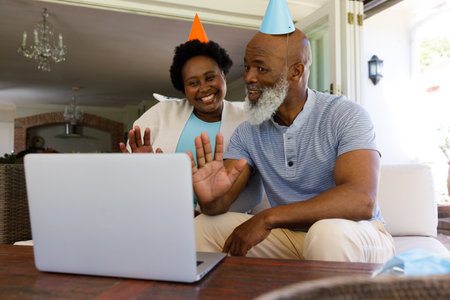 Senior african american couple spending time at home together, sitting on sofa, wearing party hats,...