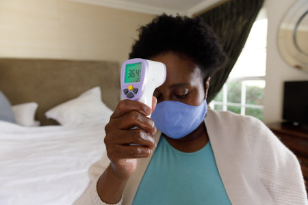 Senior african american woman spending time at home, sitting on bed, wearing face mask, taking...