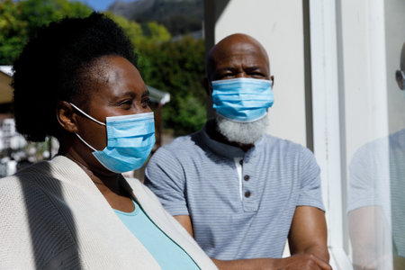 Senior african american couple spending time at home together, wearing face masks, looking through window. isolating during virus quarantine lockdown.の写真素材
