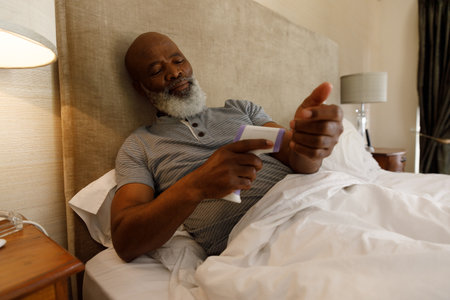 Senior african american man spending time at home, lying in bed, taking temperature on his wrist. isolating during virus quarantine lockdown.の写真素材