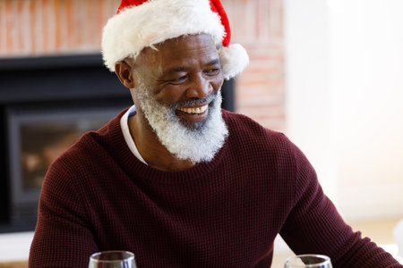 Senior african american man sitting at table, smiling and wearing santa hat. quality family time christmas celebration.の写真素材