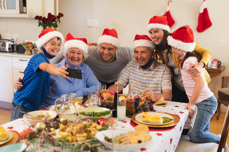 Multi generation caucasian family sitting at table for dinner together, wearing santa hats and face masks, taking a selfie and smiling. quality family time christmas celebration.の写真素材
