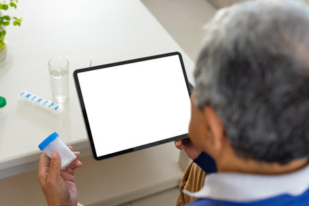 Senior biracial man sitting at table holding medicine bottle using tablet, copy space on screen. Medical services, telemedicine, senior healthcare and inclusivity concept.の写真素材