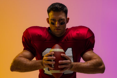 Caucasian male american football player holding ball with neon yellow and purple lighting. Sport,...
