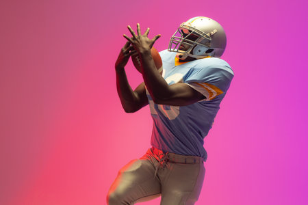 African american male american football player holding ball with neon pink lighting. Sport,...