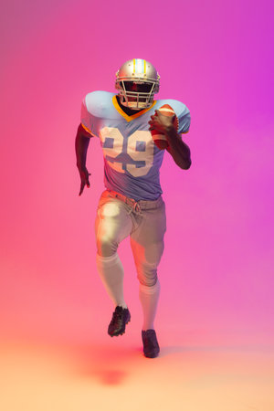 African american male american football player holding ball with neon pink lighting. Sport, movement, training and active lifestyle concept.の写真素材